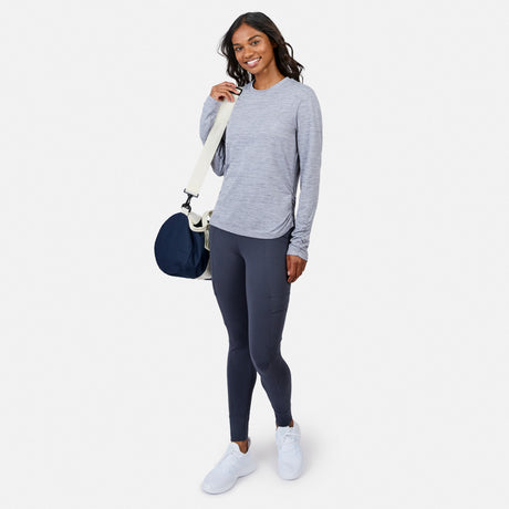 A woman smiles, wearing a Free Country Women’s Cloud Knit Crew Neck Top in light gray, dark gray leggings, and white sneakers, holding a navy and white shoulder bag against a plain white background.