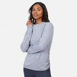 A woman wears the Free Country Women’s Cloud Knit Crew Neck Top with dark pants, standing against a white backdrop and smiling gently. This long sleeve top offers moisture-wicking comfort for all-day wear.