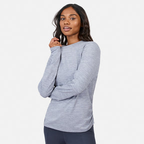 A woman wears the Free Country Women’s Cloud Knit Crew Neck Top with dark pants, standing against a white backdrop and smiling gently. This long sleeve top offers moisture-wicking comfort for all-day wear.