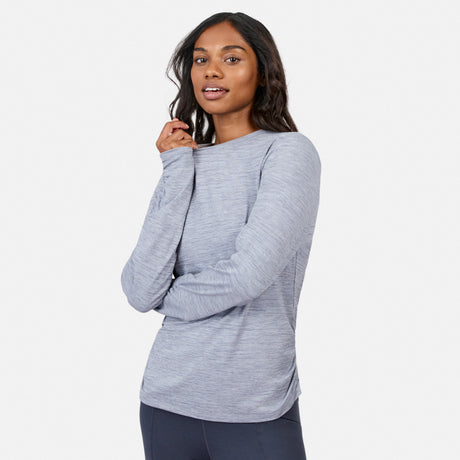 A woman wears the Free Country Women’s Cloud Knit Crew Neck Top with dark pants, standing against a white backdrop and smiling gently. This long sleeve top offers moisture-wicking comfort for all-day wear.