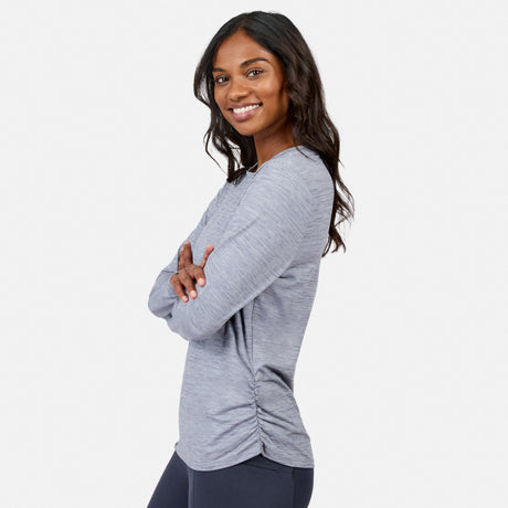 Woman with long, wavy dark hair smiles and stands sideways with arms crossed, wearing a Women’s Cloud Knit Crew Neck Top by Free Country and dark leggings against a plain white background.