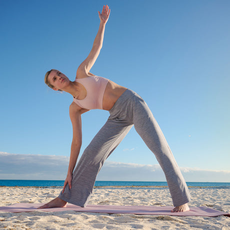 A woman practices yoga on a sandy beach in pink sports bra and Free Country Women's Cloud Knit Wide Leg Pants, stretching with one arm raised and the other touching her foot as the sea and blue sky create a calm backdrop.