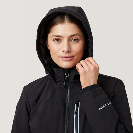 A woman wearing the Clearance Women's Thermo Super Softshell® Ski Jacket in black, featuring a hood, stands facing forward and holds the edge of the hood. The background is plain light gray.