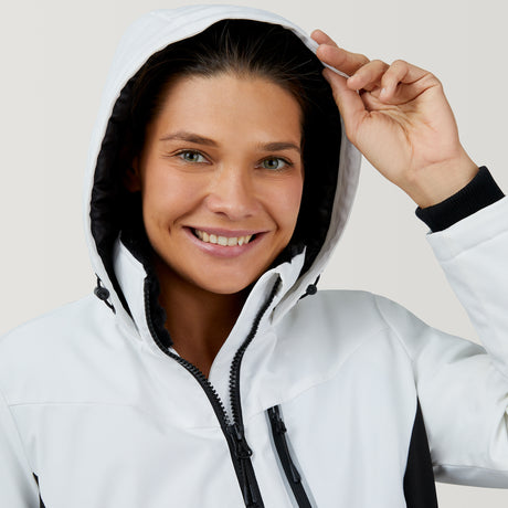 A smiling person wears the Clearance Women's Thermo Super Softshell® Ski Jacket, featuring a white, water-resistant design with black details, holding the hood’s edge and looking at the camera against a light background.