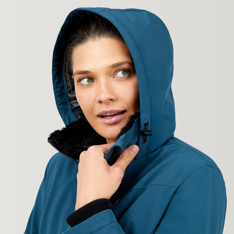 A woman models the Clearance Women's Thermo Super Softshell® Ski Jacket in blue with black fur lining, looking to the side and holding her hood against a plain light gray background.