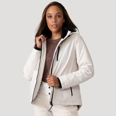 A woman models the Clearance Women's Thermo Super Softshell® Ski Jacket in white over a brown shirt, standing against a plain light background while holding the collar and zipper, looking at the camera.