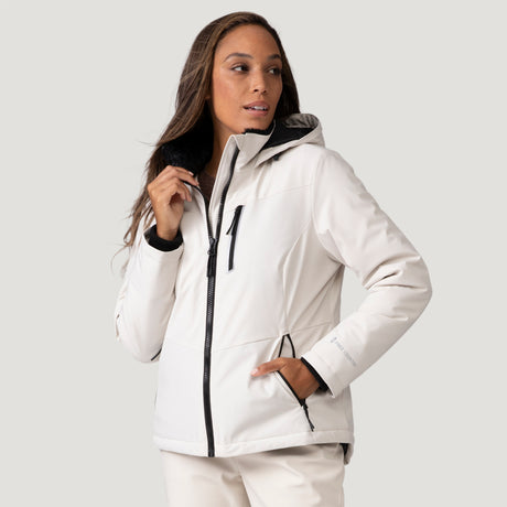 A woman wearing the Clearance Women's Thermo Super Softshell® Ski Jacket, a light-colored, water-resistant zip-up with black zippers, stands against a plain background, looking to the side with one hand on her collar and the other in her pocket.