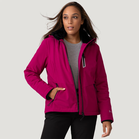 A woman stands against a light gray background wearing the Clearance Women's Thermo Super Softshell® Ski Jacket in magenta, featuring a black fur-lined hood, with black pants and one hand in her pocket.