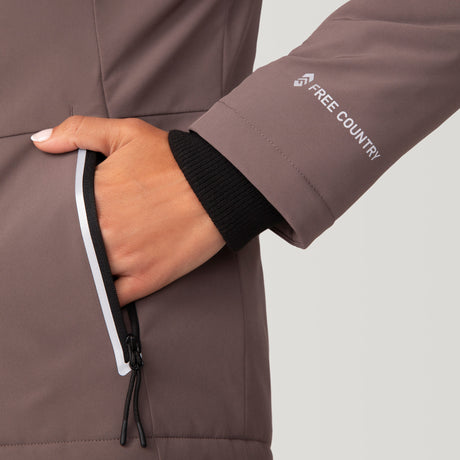 Woman wearing Free Country Women's FreeCycle® Thermo Super Softshell® II Jacket in light brown with a full front zipper opening, lower pockets, soft lining, a detachable hood with an adjustable drawcord, and water resistant and wind resistant stretch performance fabric, light grey background. Best women’s jacket for winter weather.