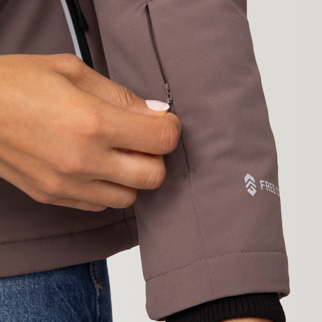 Woman wearing Free Country Women's FreeCycle® Thermo Super Softshell® II Jacket in light brown with a full front zipper opening, lower pockets, soft lining, a detachable hood with an adjustable drawcord, and water resistant and wind resistant stretch performance fabric, light grey background. Best women’s jacket for winter weather.