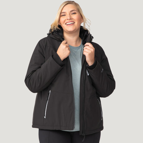 [Lola is 5'10.5" wearing a size 1X.] Woman wearing Free Country Women's Plus Size FreeCycle® Thermo Super Softshell® II Jacket in black with a full front zipper opening, lower pockets, soft lining, a detachable hood with an adjustable drawcord, and water resistant and wind resistant stretch performance fabric, light grey background. Best women’s jacket for winter weather.