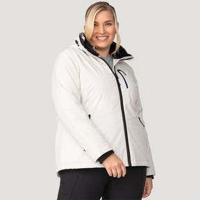 A smiling woman with blonde hair wears the Women's Plus Size FreeCycle® Thermo Super Softshell® II Jacket by Clearance in Oyster, featuring black zippers and REPREVE recycled insulation, paired with black pants, standing against a plain light gray background.