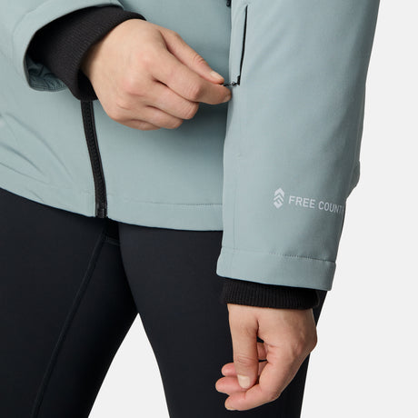 A woman zips the sleeve pocket of a light blue Women's Plus Size FreeCycle® Thermo Super Softshell® II Jacket by Free Country, featuring water-resistant fabric and logo, partially open to show a black inner layer and paired with black pants.