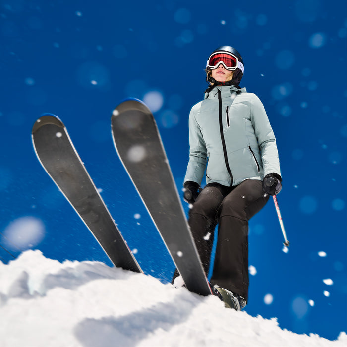 A skier stands on a snowy slope wearing a light sage green ski jacket, black ski pants, black gloves, and a black helmet with red-tinted goggles. Two skis are visible in the snow against a bright blue sky with falling snow.
