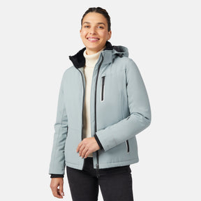 A woman wears the Free Country Women's FreeCycle® Thermo Super Softshell® II Jacket in light blue over a white turtleneck and black pants, smiling against a plain white background.