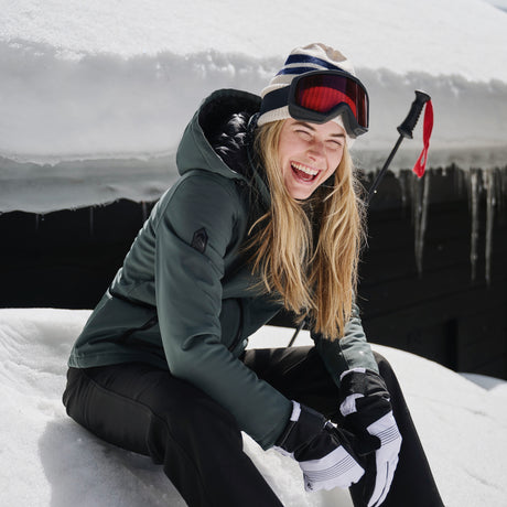 Wearing the Free Country Women's Thermo Super Softshell® Long Jacket with quilted lining, a woman sits on snow, smiling, as ski poles rest behind her and a building with icicles appears in the background.