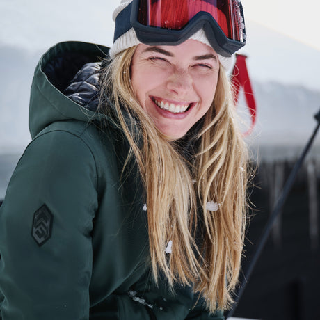 A smiling woman with long blonde hair wears the Free Country Women's Thermo Super Softshell® Long Jacket in green, paired with a white beanie and red ski goggles on her forehead. Snow in the background hints at a wintry outdoor scene.