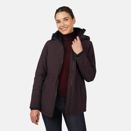 A woman smiles, wearing the Free Country Women's Thermo Super Softshell® Long Jacket in dark purple over a burgundy turtleneck and dark jeans, with one hand on her collar, against a plain white background.