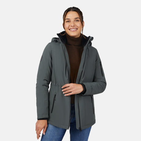 A smiling woman wears the Free Country Women's Thermo Super Softshell® Long Jacket in dark green, featuring a quilted interior lining and hood, over a brown turtleneck sweater and blue jeans, standing against a white background.