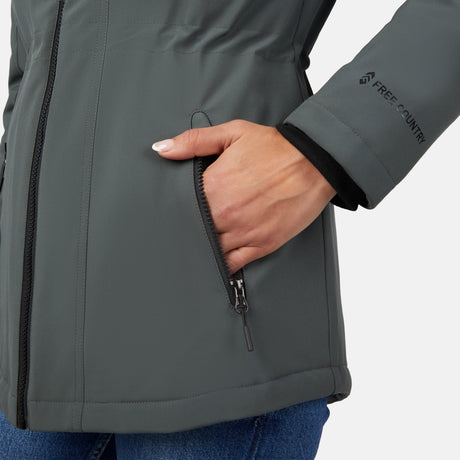 A woman in blue jeans unzips a side pocket on the Free Country Women's Thermo Super Softshell® Long Jacket. The gray, water-resistant jacket has the brand logo near the wrist and a quilted interior lining for added warmth.