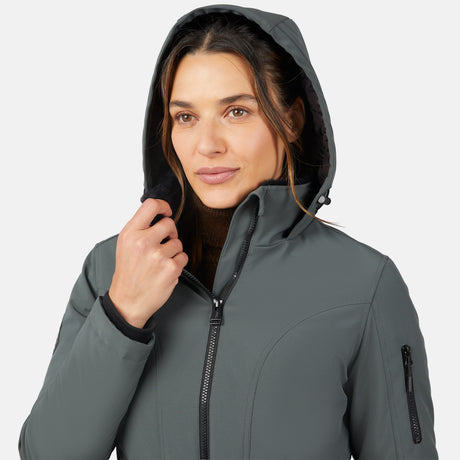 A woman in the Free Country Women's Thermo Super Softshell® Long Jacket stands against a plain white background, holding her hood. The water-resistant gray jacket has a front zipper, zippered sleeve pocket, and quilted interior lining.