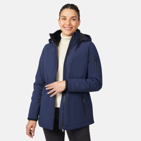 A woman smiles against a plain background, wearing the Free Country Women's Thermo Super Softshell® Long Jacket in navy blue over a white turtleneck and black pants, with one hand on the jacket’s zipper. The jacket features water resistance and quilted lining.