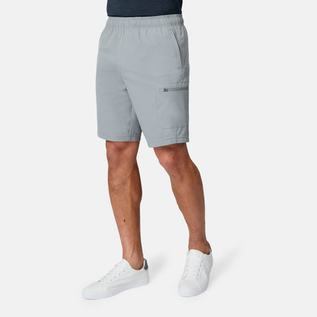 A man wears Free Country Men's Tech Stretch Short II in light gray with an elastic waistband, side pocket, and moisture-wicking comfort, paired with white sneakers against a plain white background. His upper body is not visible.