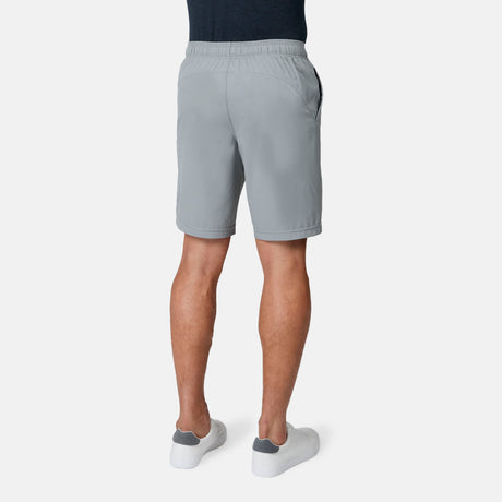 A man is seen from the back wearing Free Country Men's Tech Stretch Short II in light gray, paired with a dark shirt and white sneakers, standing against a plain white background.