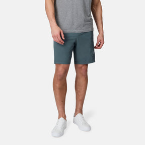 A person wearing a gray t-shirt, Free Country Men's Tech Stretch Short II in teal, and white sneakers stands against a plain white background with their arms relaxed by their sides.