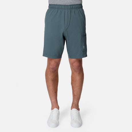 A person showcases only the lower half, wearing Free Country Men's Tech Stretch Short II in teal and white sneakers, with a gray shirt tucked in, standing against a plain white background.