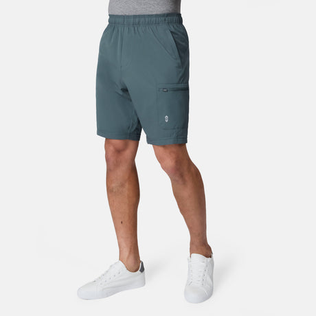 A man wears Free Country Men's Tech Stretch Short II in teal, featuring a stretch weave, paired with white sneakers against a white background.