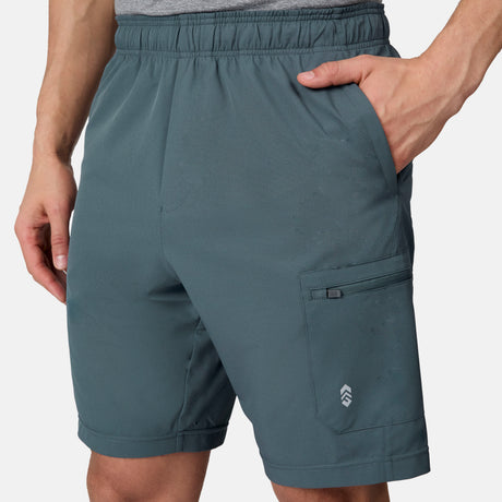 A person wears Free Country Men's Tech Stretch Short II in teal, featuring an elastic waistband, side pockets, and a right thigh zip pocket. Paired with a gray shirt, the moisture-wicking fabric keeps them cool as their right hand rests in the pocket.