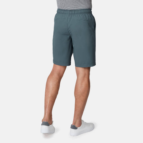 A person wearing Free Country Men's Tech Stretch Short II in teal and a gray t-shirt, paired with white sneakers, is shown from the back against a plain white background.
