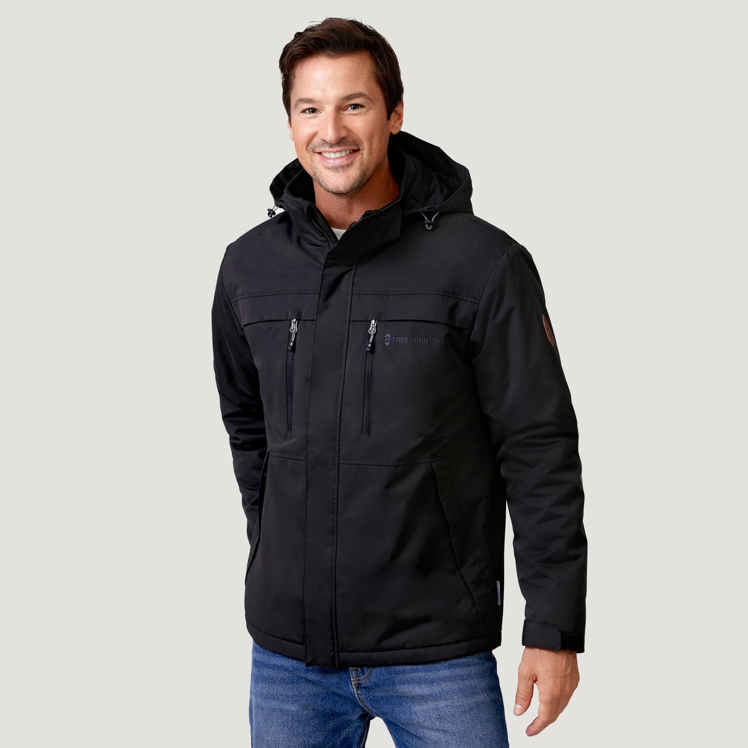 A man smiles while wearing a black Men’s Wild Land Mid Weight Jacket with zippered chest pockets and blue jeans.
