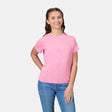 A young woman smiles against a white background, wearing a pink Free Country Girls' Microtech® Crew Neck Shirt and blue jeans, with one hand on her hip and the other relaxed by her side.