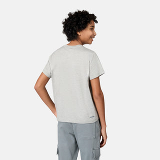 A person with short curly hair, wearing the Free Country Boys' Microtech® Graphic Crew Neck Shirt in light gray and paired with gray cargo pants, stands with one hand on their hip, facing away from the camera against a plain light background. The moisture-wicking Microtech® fabric of the shirt ensures comfort throughout the day.