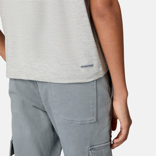 A person is shown from the waist down wearing gray cargo pants and a light gray Boys' Microtech® Graphic Crew Neck Shirt by Free Country, made from moisture-wicking Microtech Chill fabric with UPF 50+ protection and featuring a small MICRO-TECH label near the hem. One arm is relaxed by their side.