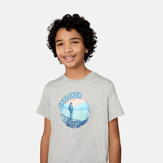 A smiling child with curly hair is wearing the Free Country Boys' Microtech® Graphic Crew Neck Shirt in light gray, featuring a graphic of a person standing on rocks with mountains and the word "EXPLORER" above. Made from soft, moisture-wicking Microtech Chill fabric for all-day comfort. Plain white background.