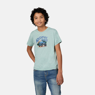 A smiling boy with curly hair wears a light blue Free Country Boys' Microtech® Graphic Crew Neck Shirt featuring an Adventure Awaits car graphic, crafted from soft Microtech Chill fabric for comfort and moisture-wicking performance, paired with blue jeans and standing against a plain white background.