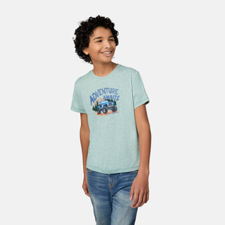 Smiling boy with curly hair wears a Free Country Boys' Microtech® Graphic Crew Neck Shirt featuring "Adventure Awaits" and an off-road vehicle, paired with blue jeans, standing against a plain light background.