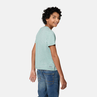 A smiling boy with curly hair wears the Free Country Boys' Microtech® Graphic Crew Neck Shirt in light green and blue jeans, looking over his shoulder against a plain white background.