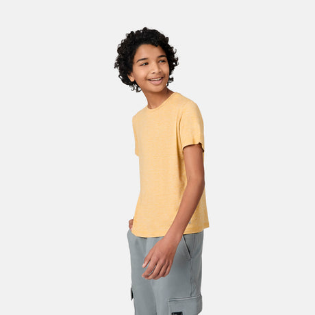 A smiling young person with curly hair wears a Free Country Boys' Microtech® Crew Neck Shirt and light gray cargo shorts, standing against a plain light background.