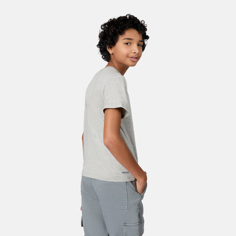 A young person with curly hair wears the Free Country Boys' Microtech® Crew Neck Shirt and gray pants, standing sideways with one hand in their pocket and smiling over their shoulder against a plain white background.