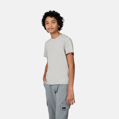 A young person with curly hair smiles with one hand in their pocket, wearing a light gray Free Country Boys' Microtech® Crew Neck Shirt and gray cargo pants, standing against a plain white background.