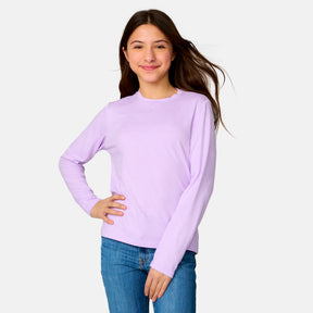 A young woman with long brown hair smiles confidently with one hand on her hip, showcasing the Clearance Girls' Microtech® Long Sleeve Shirt in light purple paired with blue jeans against a simple, light background.