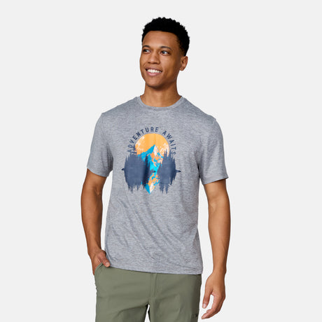 A smiling man with short, curly hair wears the Clearance Men's Super Soft Short Sleeve Crew Tee - 2 Pack in gray, featuring mountains, trees, a sunset, and "Adventure Awaits," paired with olive-green pants and one hand in his pocket.