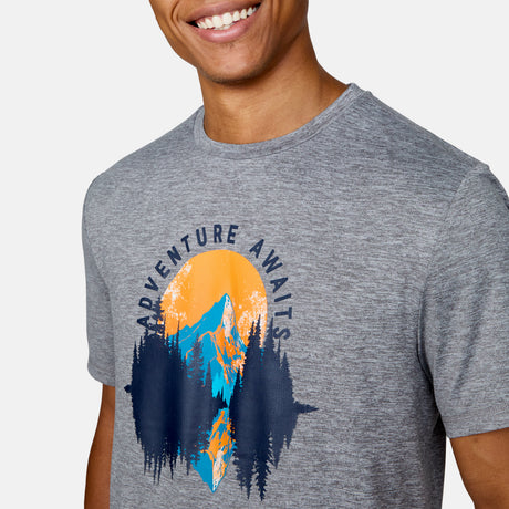 A person in a Clearance Men's Super Soft Short Sleeve Crew Tee - 2 Pack, gray with a vibrant mountains, trees, and sun graphic and "ADVENTURE AWAITS" text. This moisture-wicking tee keeps them smiling and comfy all day.