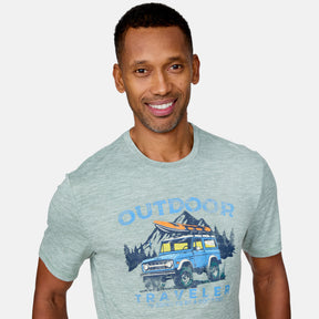 A smiling man wears a light green Clearance Men's Super Soft Short Sleeve Crew Tee featuring a yellow SUV, pine trees, and mountains. This 2-pack OUTDOOR TRAVELER shirt offers UPF 50+ protection. Plain white background.