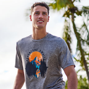 A young man smiles outdoors in a Clearance Men's Super Soft Short Sleeve Crew Tee—2 Pack, featuring moisture-wicking fabric and a mountain sunset Adventure Awaits graphic, with palm trees and a bright sky behind him.