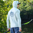 A man wearing the Free Country Men's Microtech® Hooded Graphic Sunshirt with UPF 50+ holds a green-bladed paddle near water. The shirt says "Lake Time Canoe & Kayak" with pine trees, and there are trees in the background.